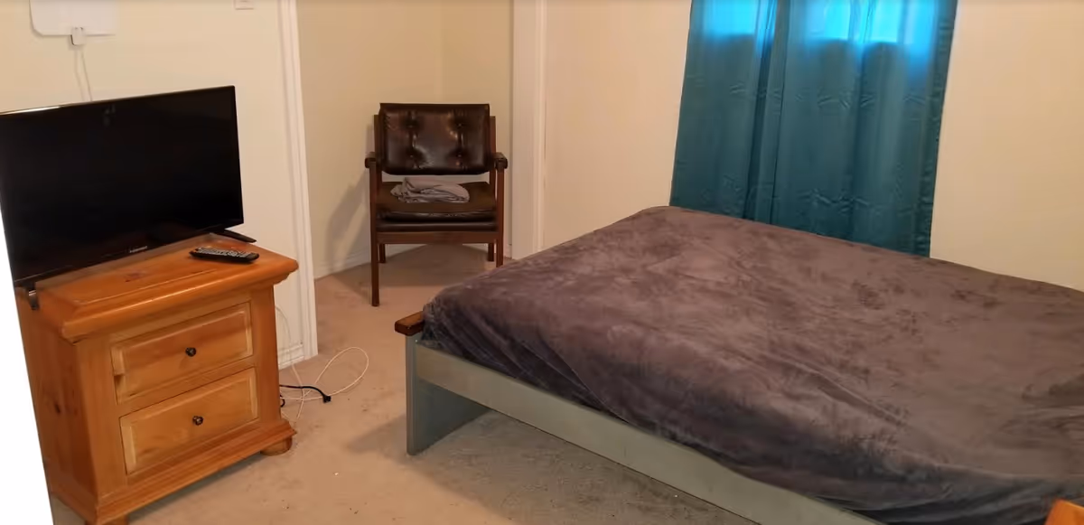 Bedroom in Magda's Adult Care, featuring bed, TV, dresser, chair, and teal curtains.