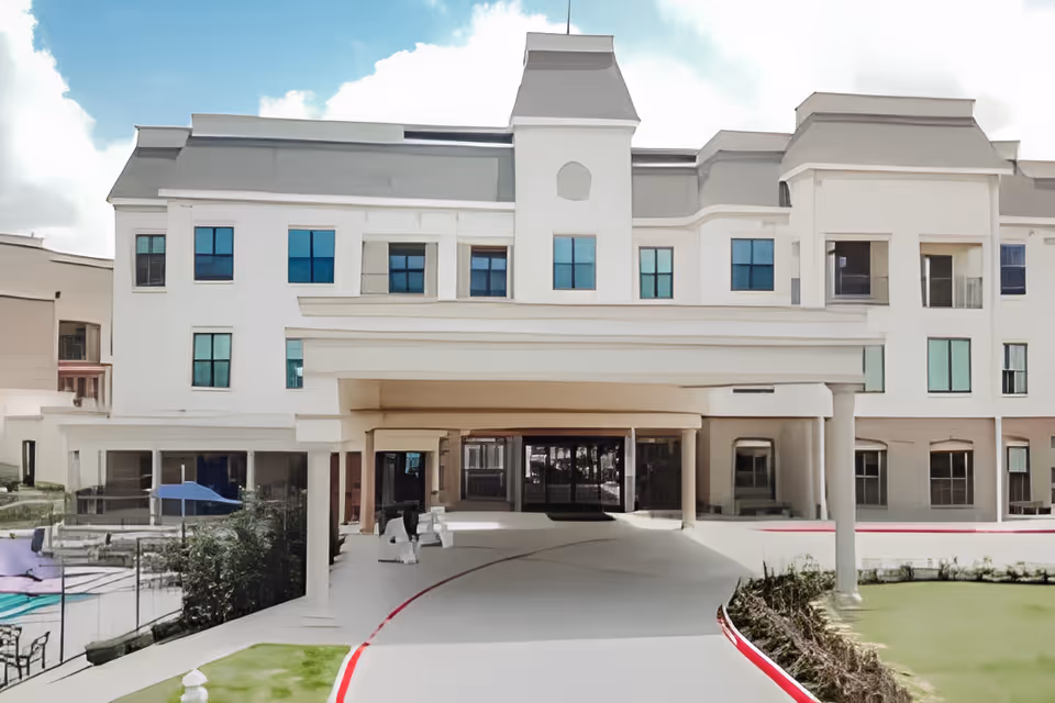 Front exterior view of a multi-story senior living facility building with a covered entrance driveway, large windows, and a landscaped area with grass and bushes.