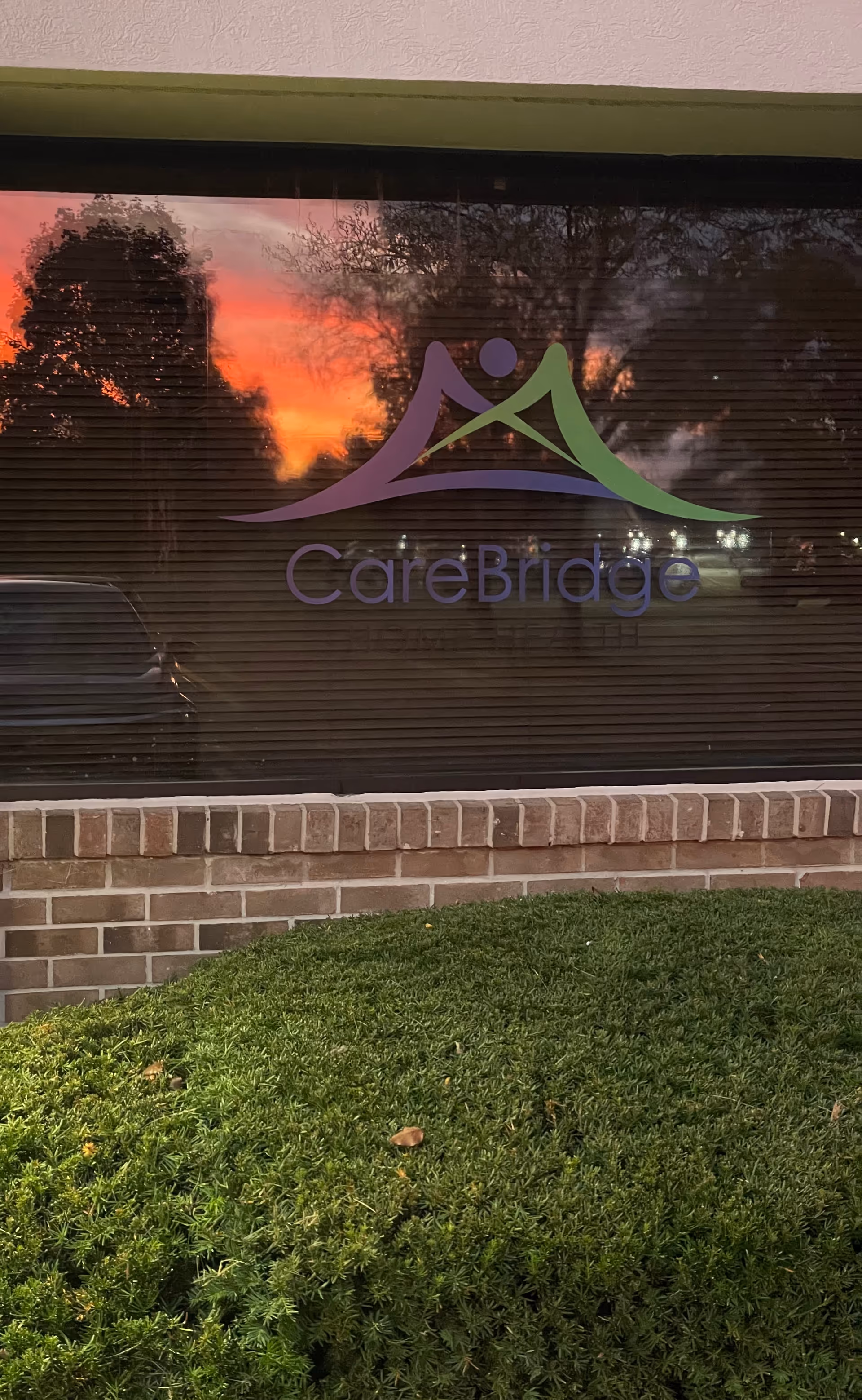 Glass window displays CareBridge logo with sunset sky reflection, brick wall, and manicured bushes outside.