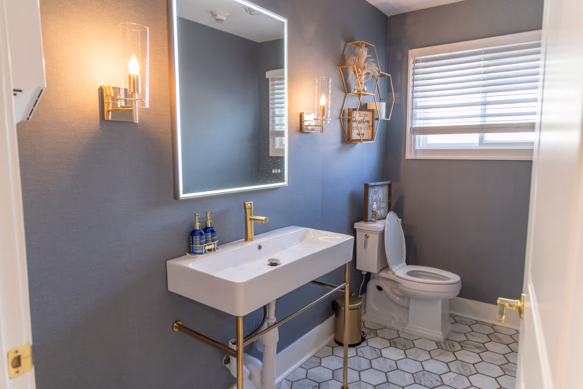 Blue-walled bathroom with a white sink, mirror, toilet, hex tile floor, and window.