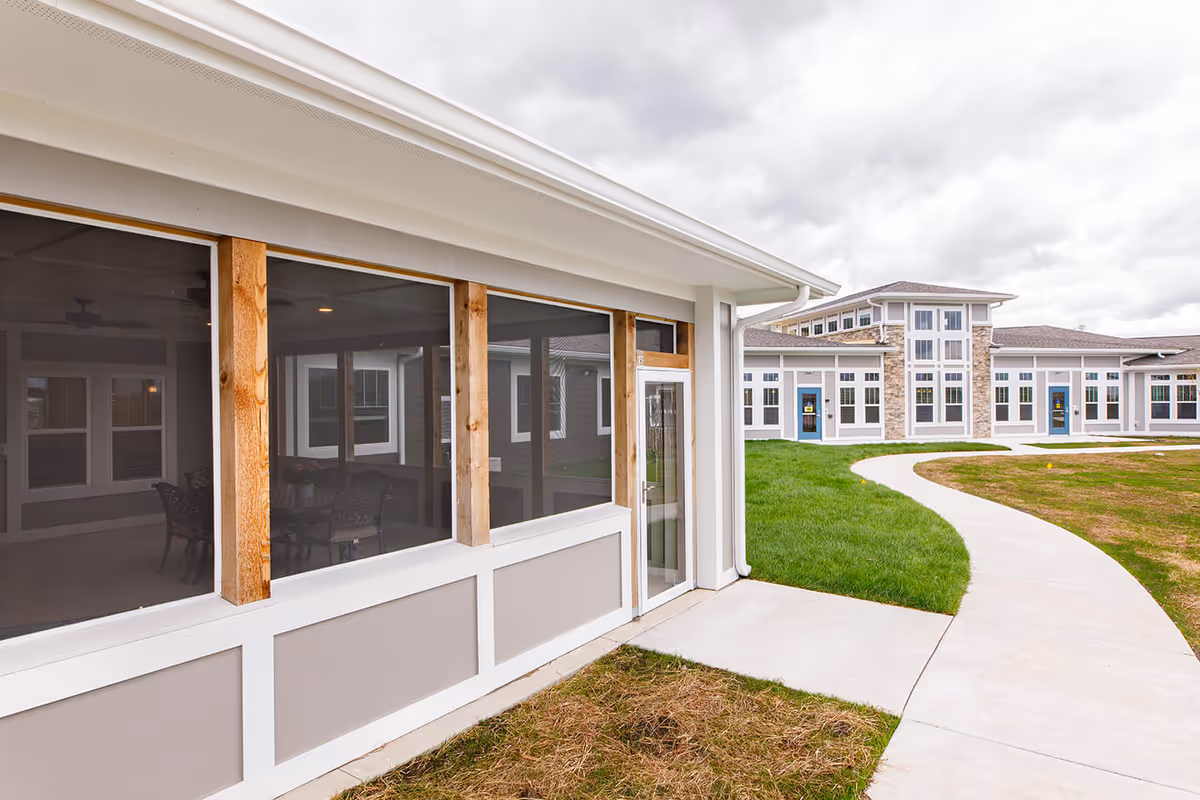 Exterior view of a modern assisted living building with a screened porch, curved walkway, and lawns under a cloudy sky.