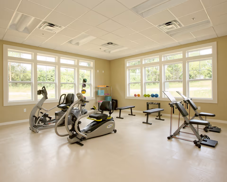 Bright fitness room with recumbent exercise machines, benches, small weights and large windows overlooking greenery.