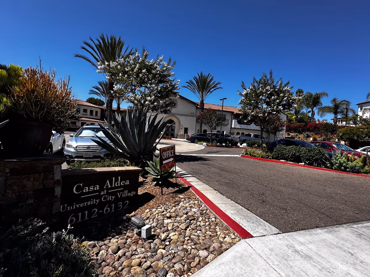 Entrance and driveway of Casa Aldea at University City Village showing the property sign, landscaping, parked cars, and the building under a clear blue sky.