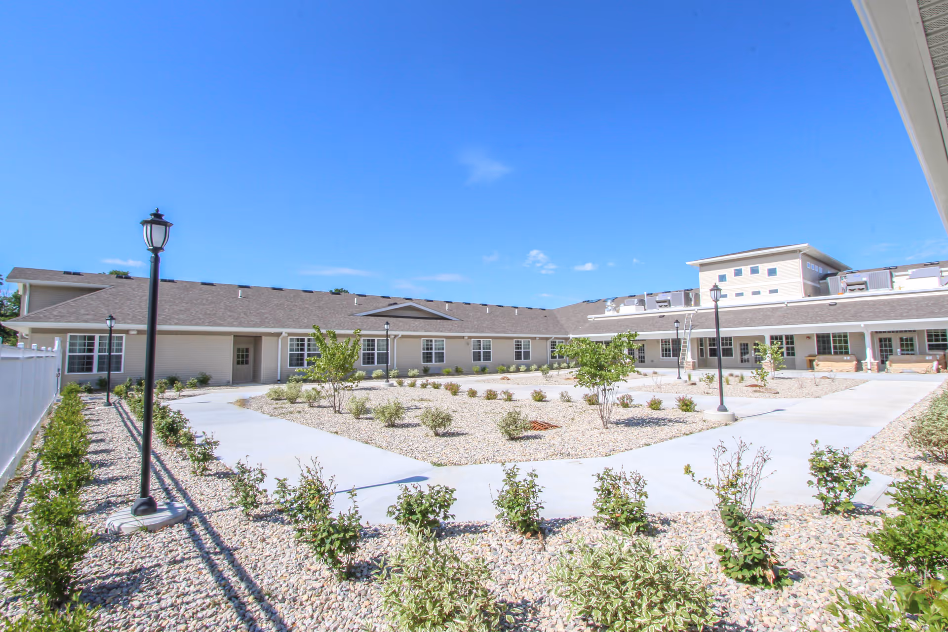 Outdoor courtyard area of Lincolnshire Place - Memory Care & Assisted Living with paved walkways, small shrubs, young trees, and lamp posts under a clear blue sky.