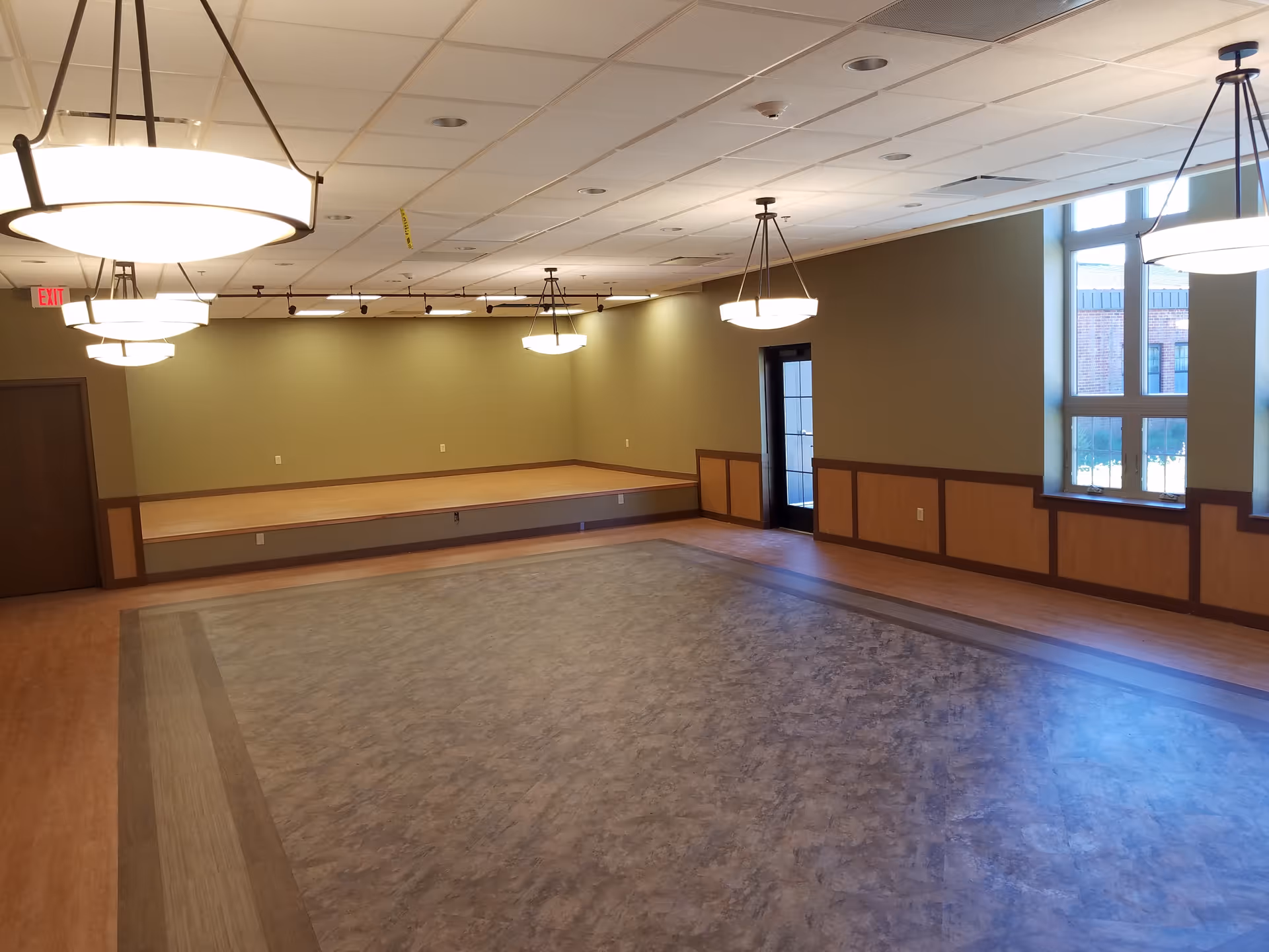Interior multipurpose room in a senior facility with stage, lighting fixtures, and windows.