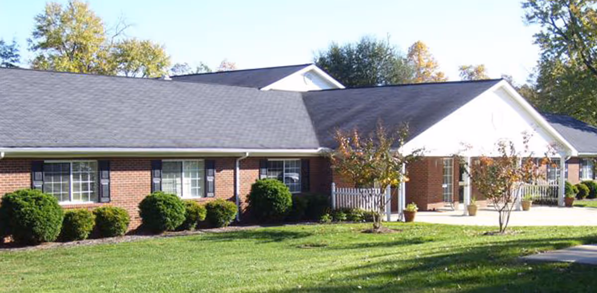 Single-story brick building with a gabled white entrance, shuttered windows, manicured shrubs and a grassy lawn.