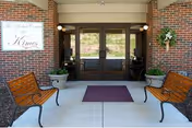 Brick building entrance with glass doors, benches, potted plants, and a maroon doormat.