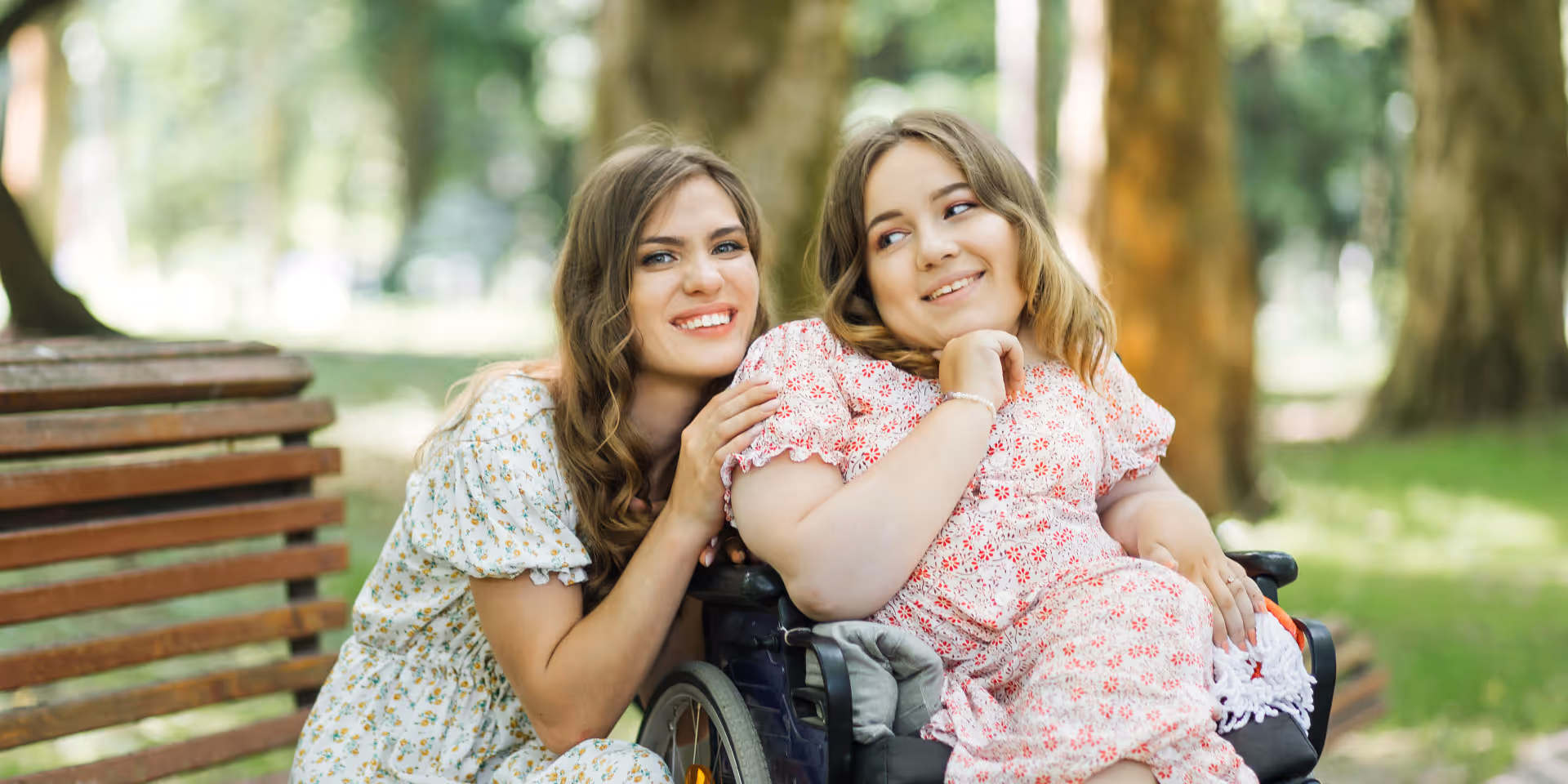 Two smiling women sit on a park bench outdoors among trees.