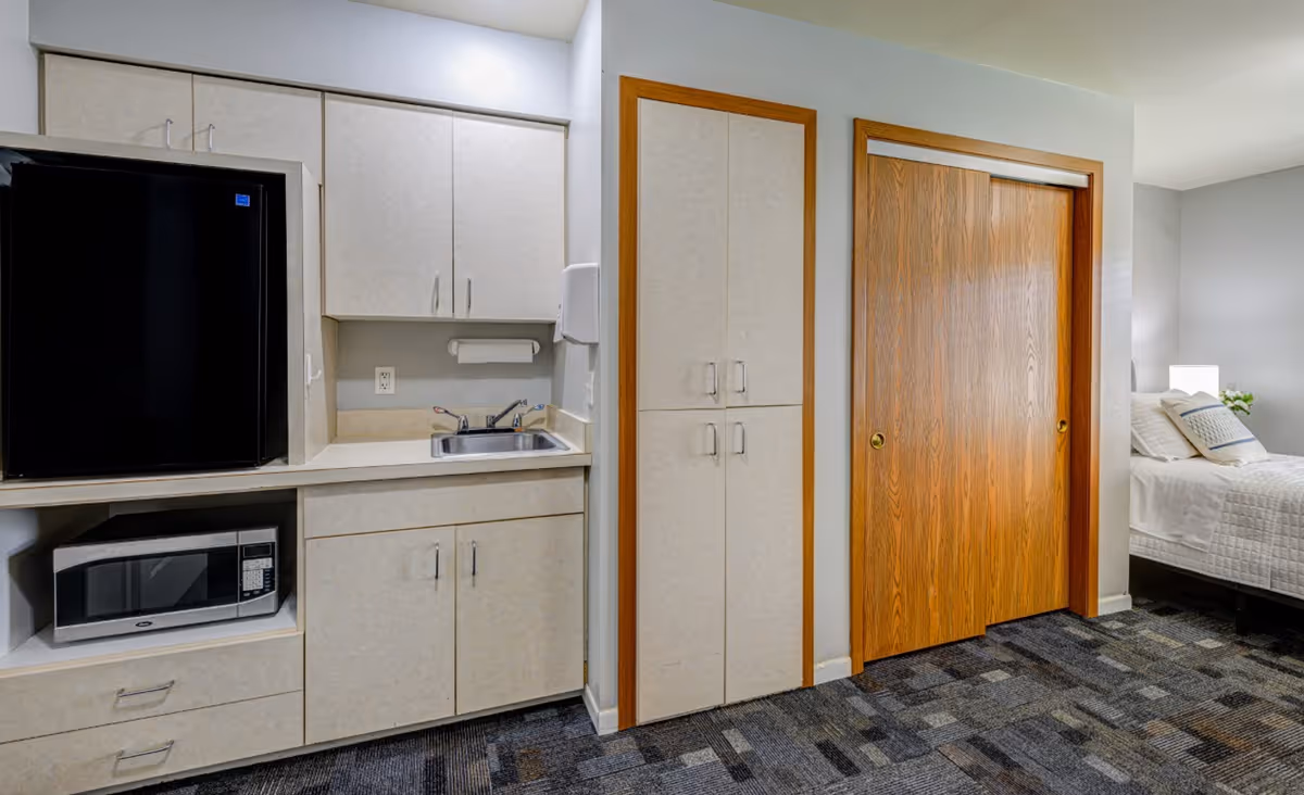 Interior view of a senior living facility room showing a kitchenette area with a black mini refrigerator, microwave, sink, and cabinets. To the right, there is a wooden sliding door closet and a bed with white bedding and pillows partially visible.