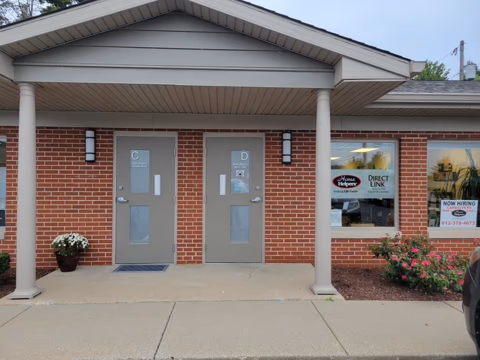 Brick building entrance with two gray doors under a covered walkway and flowering plants.