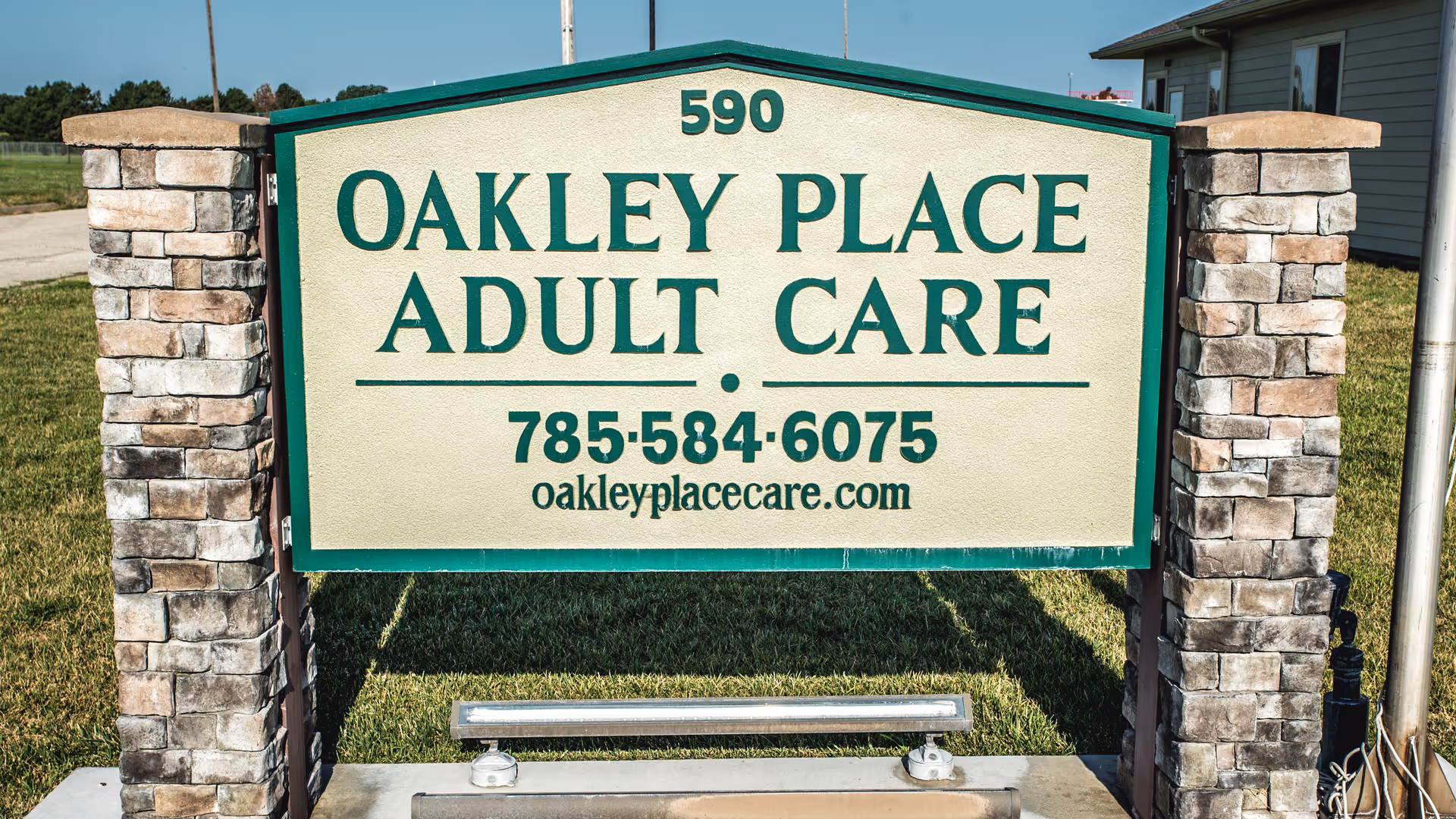 Freestanding sign reading 'OAKLEY PLACE ADULT CARE' with phone number and website between stone pillars on a grassy lawn.