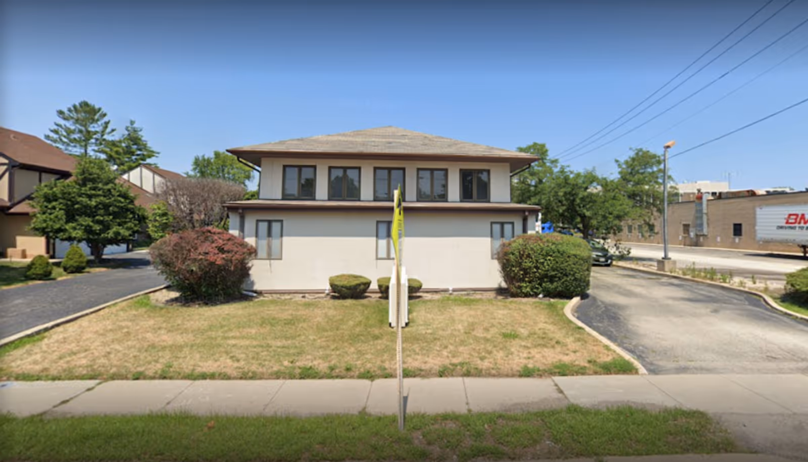 Two-story beige building with windows, trimmed shrubs, and a driveway seen from the sidewalk.
