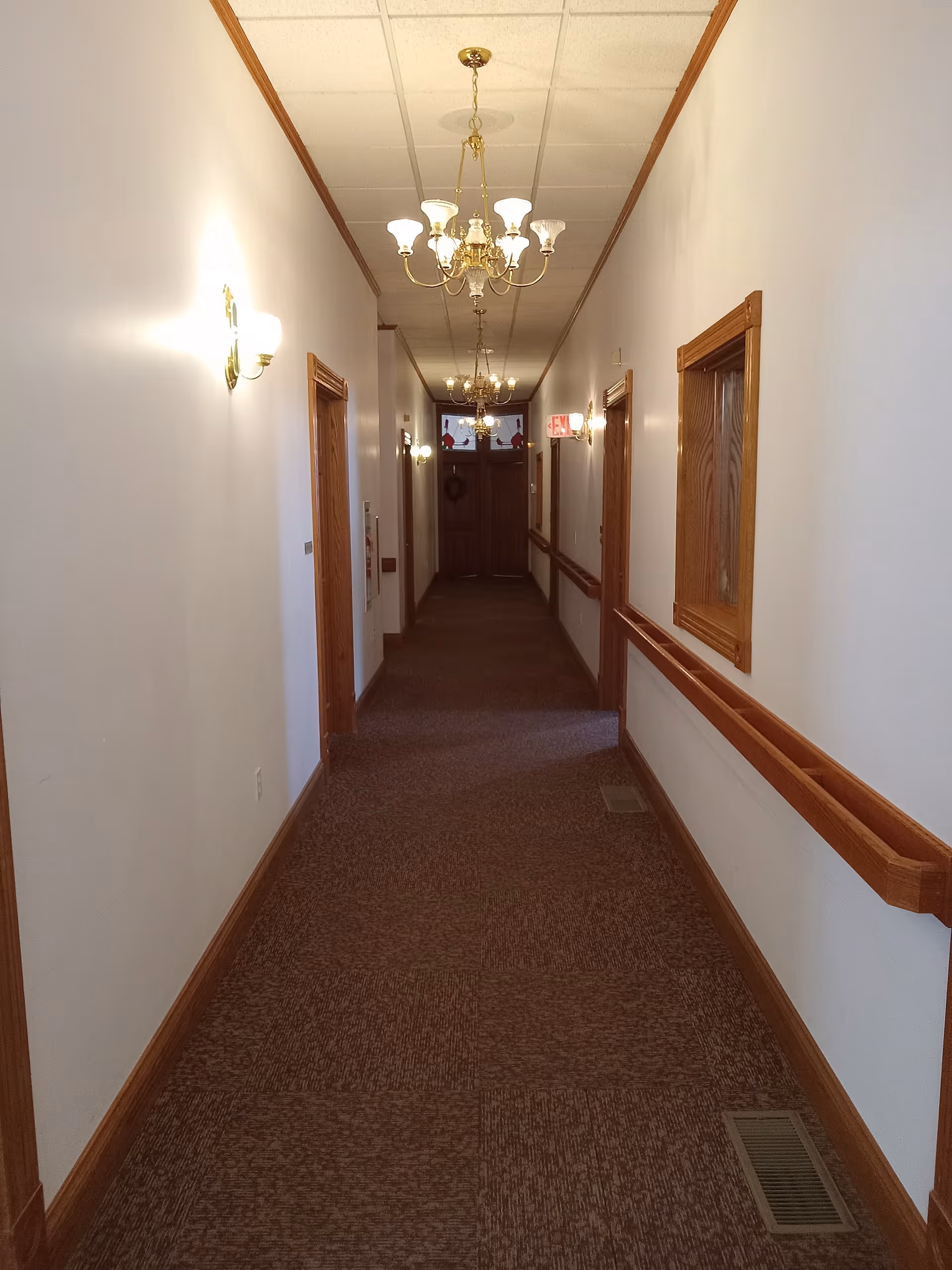 Long hallway in a senior living facility with carpet, wooden railings, and chandeliers.