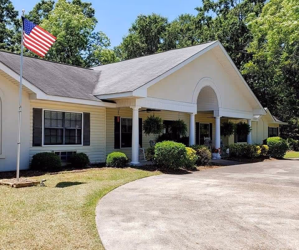 Exterior view of a single-story building with white siding and a gray roof, featuring a covered entrance supported by white columns. There are bushes and hanging plants around the entrance, an American flag on a flagpole to the left, and trees in the background under a clear blue sky.