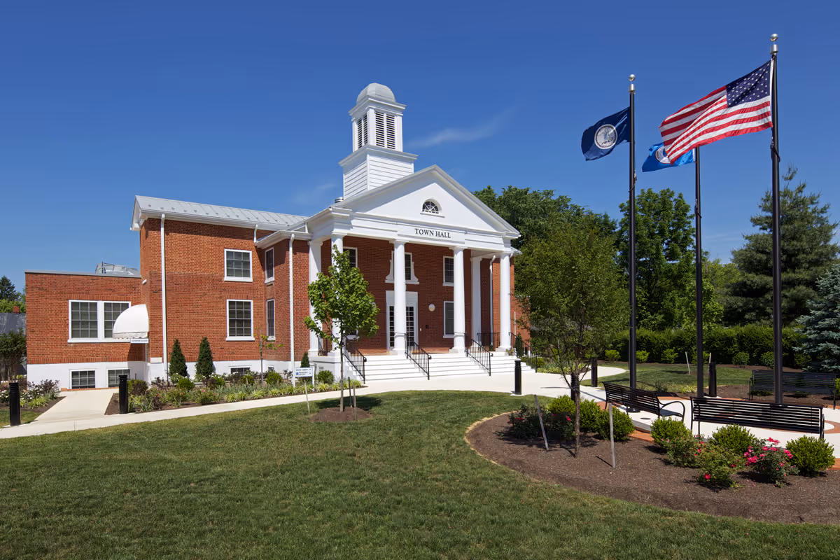 Brick town hall-style building with white columns, flags, and manicured lawn.