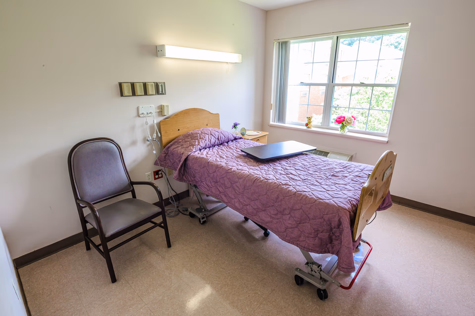 Bedroom in senior facility featuring a hospital-style bed, chair, and a sunny window.