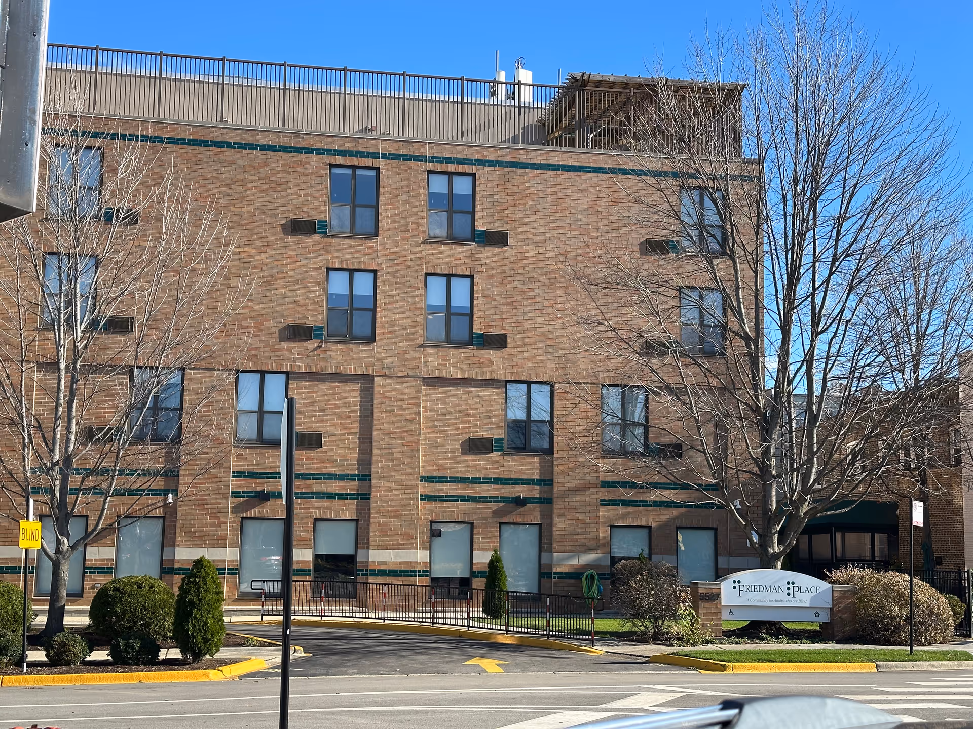 Exterior view of a multi-story brick building with several windows, leafless trees, and a sign in front that reads 'Friedman Place, A Community for Adults who are Blind'. The sky is clear and blue.