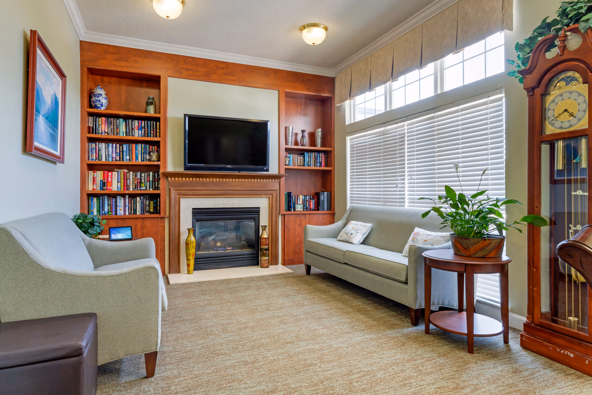 A cozy living room with a beige carpet, two light gray sofas, a wooden side table with a green plant, a large window with blinds and valance, a wooden grandfather clock, a fireplace with a TV mounted above it, and built-in wooden bookshelves filled with books and decorative items.