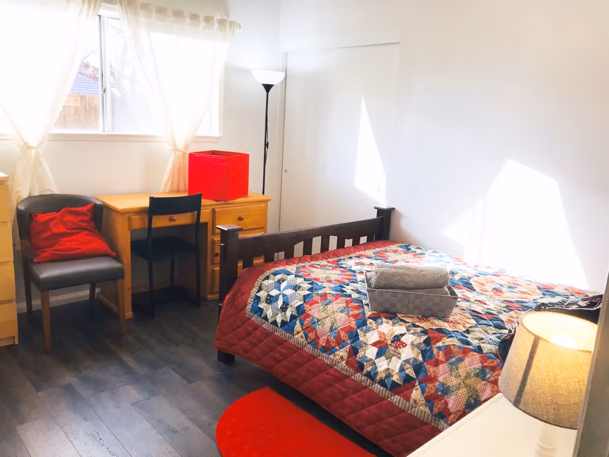 A cozy bedroom with a quilted bed, desk, chair, dresser, and sunlit window.
