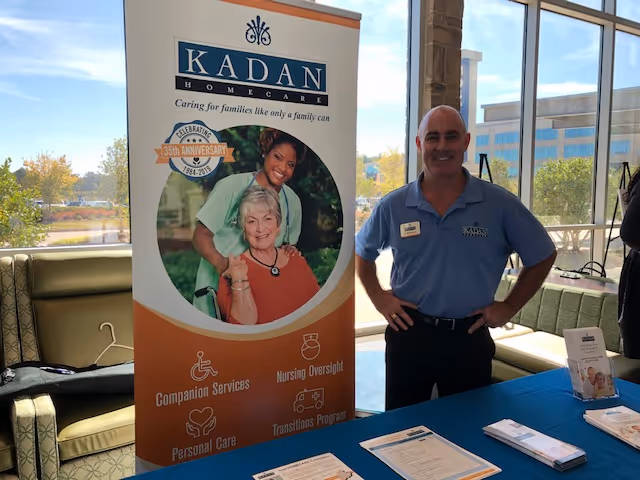 A staff member in a blue polo stands near a Kadan Homecare banner indoors.