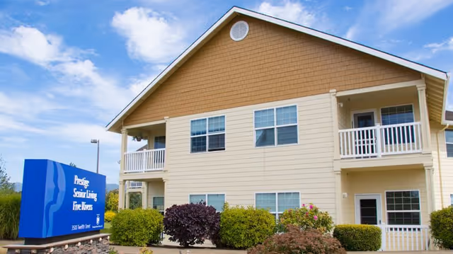 Exterior view of a two-story beige building with white trim and balconies, surrounded by bushes and landscaping under a partly cloudy sky. A blue sign in front reads 'Prestige Senior Living Five Rivers' with the address 3501 Main Street.