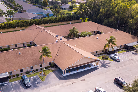 Aerial view of a single-story assisted living building with a central covered entrance, parking lot, and palm trees.
