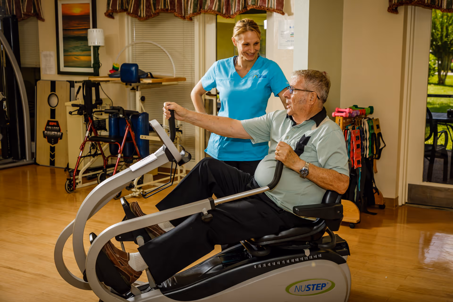 An elderly man exercises on a NuStep recumbent cross trainer machine in a rehabilitation or fitness room while a female healthcare worker in blue scrubs stands beside him, smiling and offering support. The room has wooden floors, exercise equipment, and a window with a view of greenery outside.