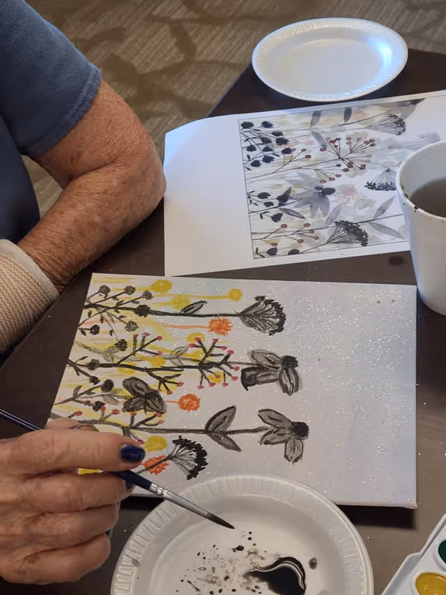 An elderly person painting a floral design on a canvas using a paintbrush and black paint. The canvas features flowers with black, yellow, and orange colors. There is a paper plate with black paint and a cup of water on the table, along with a printed reference image of a similar floral design.