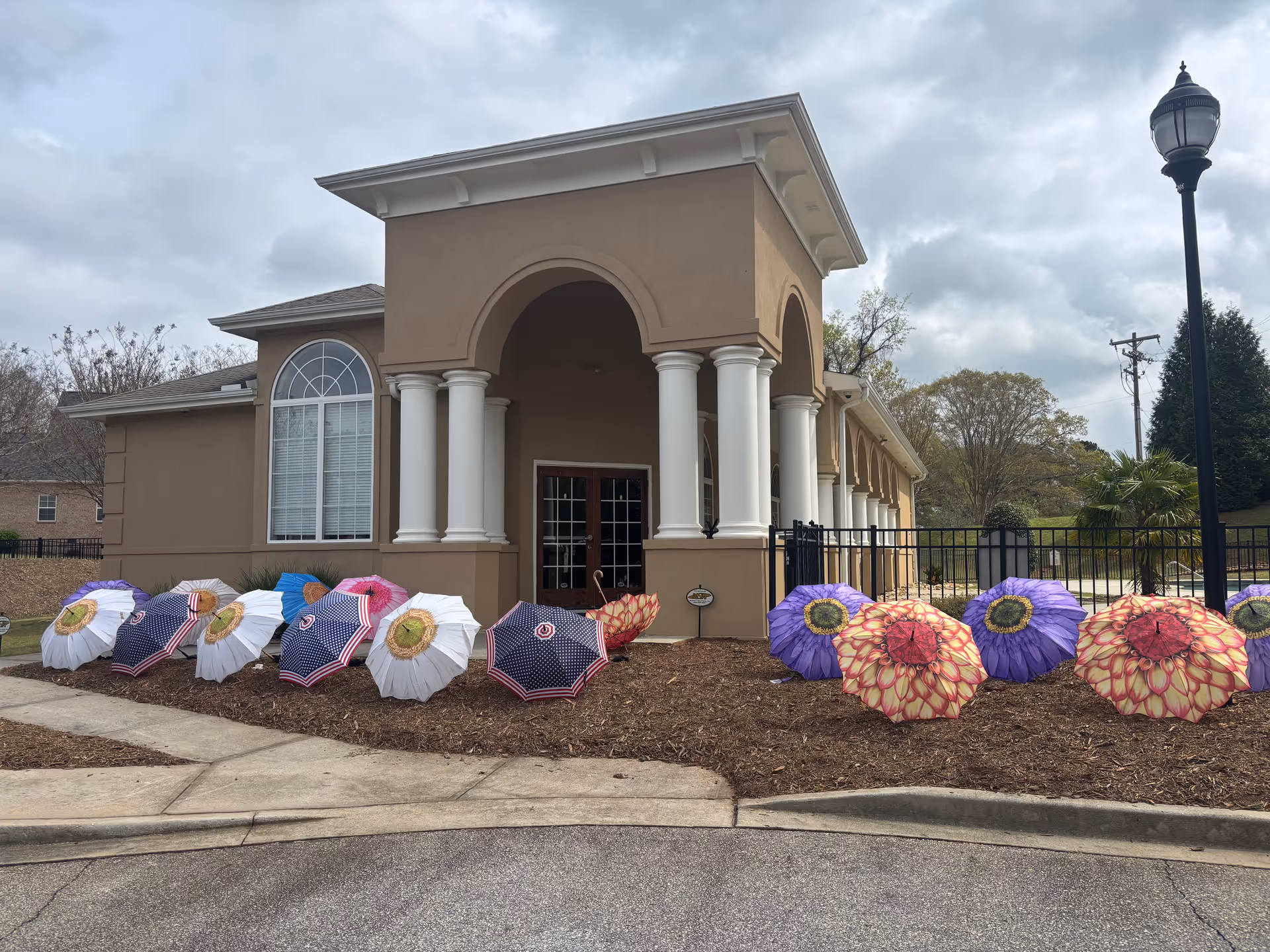 A tan senior care building exterior with arched entrance, columns, and colorful umbrellas lining the walkway.