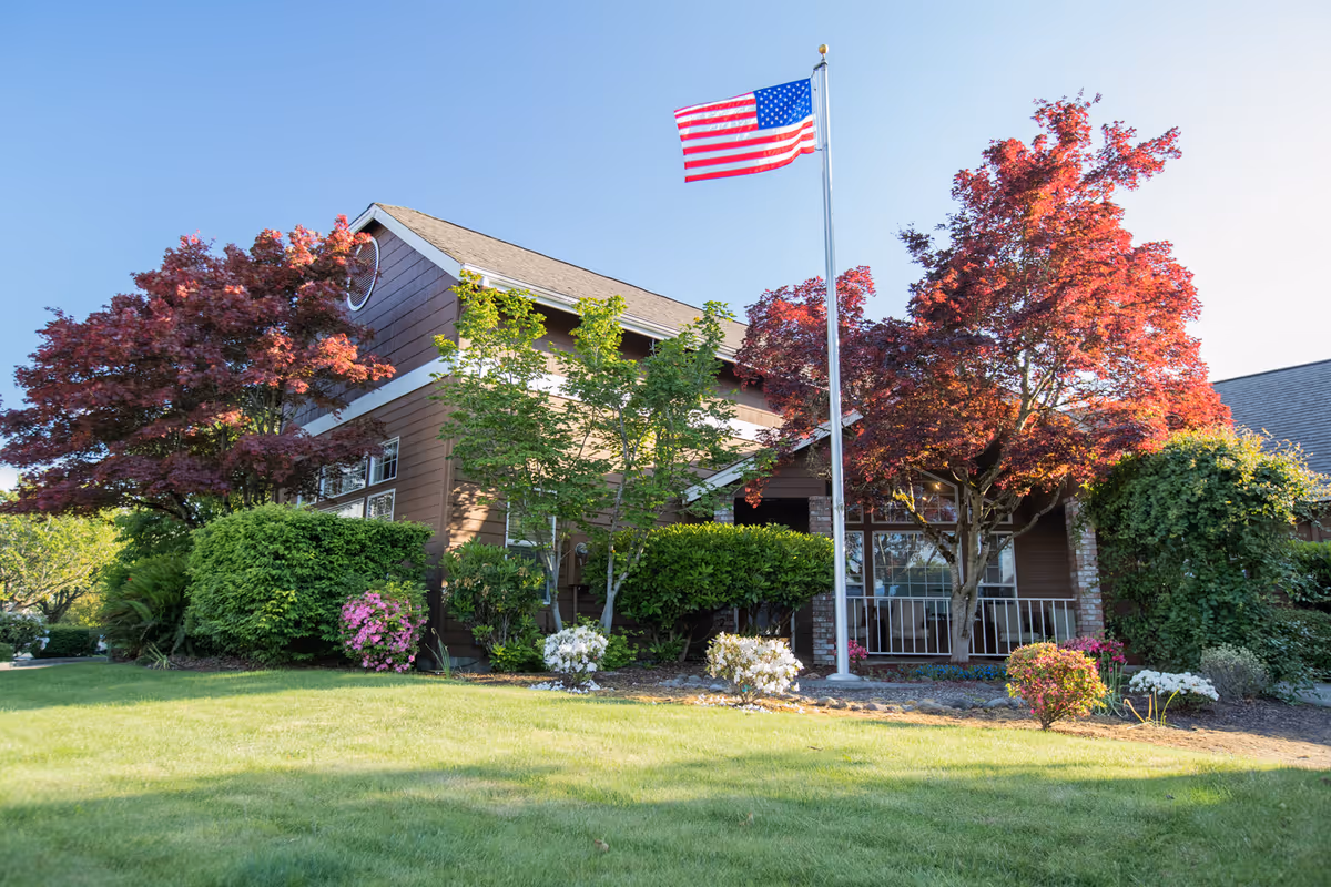 Exterior view of a brown building with a sloped roof surrounded by green and red-leaved trees and bushes. An American flag is flying on a flagpole in front of the building. The sky is clear and blue.