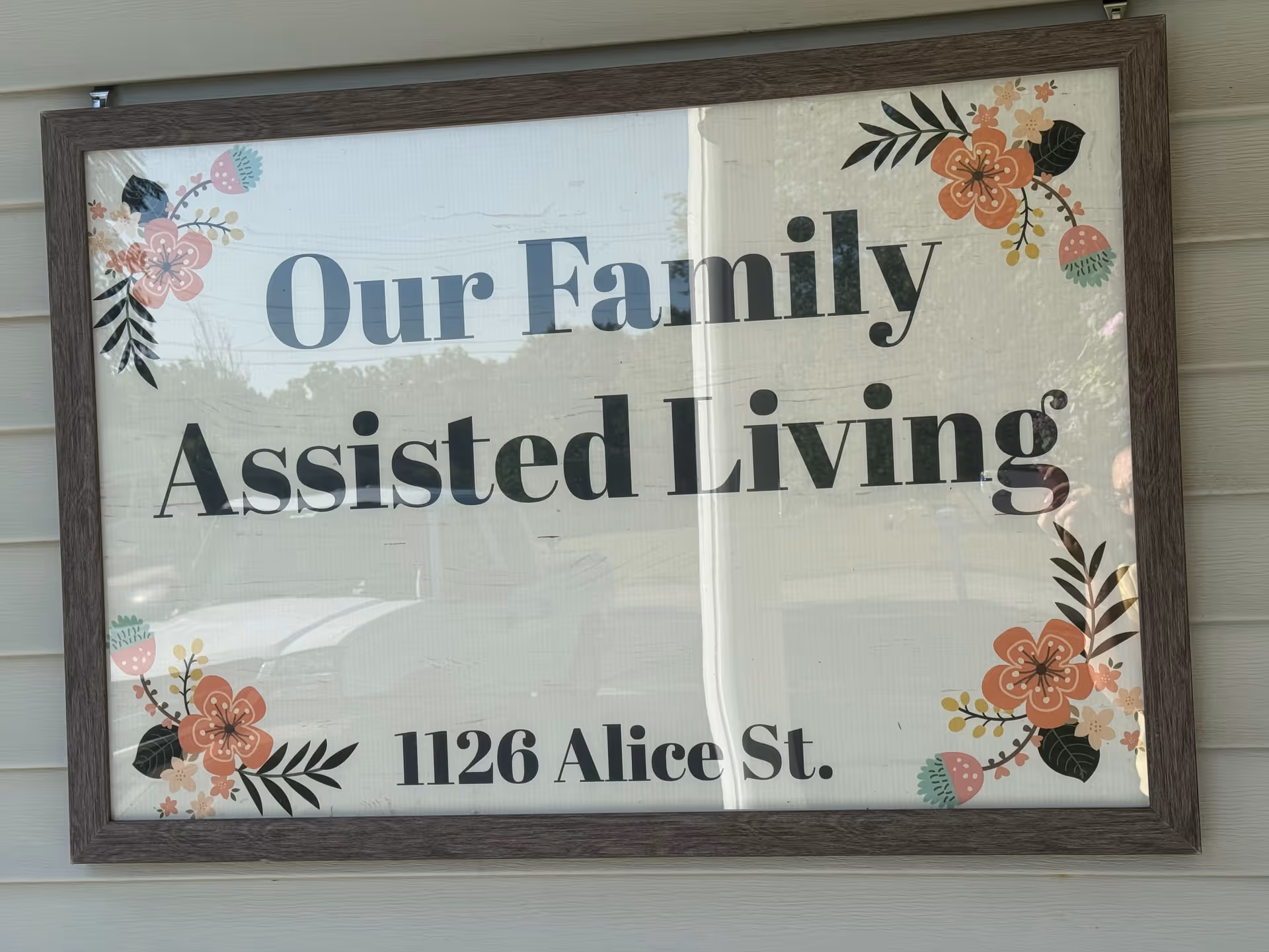 Framed sign on exterior wall reading Our Family Assisted Living with floral corners.