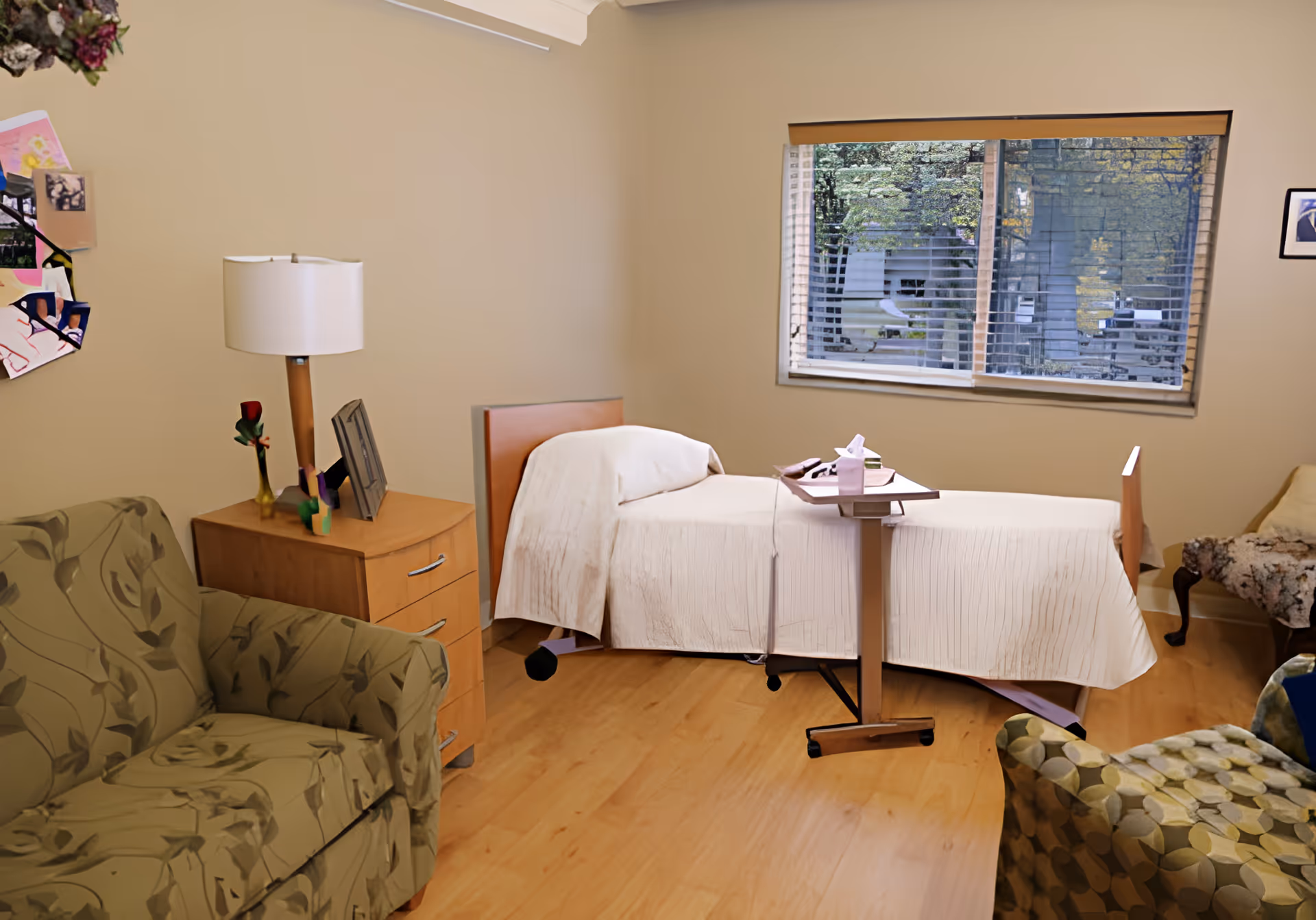 A cozy room with a single bed covered in a white blanket, a wooden bedside table with a lamp and a single red rose in a vase, a green patterned armchair, and a window with blinds showing an outdoor view with trees and buildings.