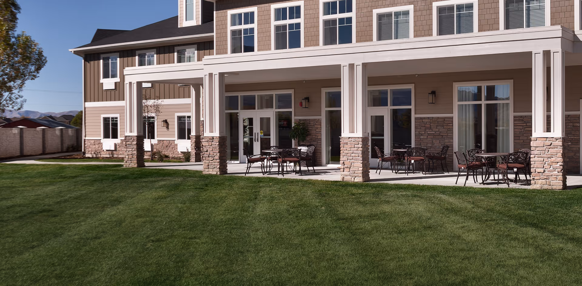 Front exterior of a senior living building with a covered patio, outdoor tables and chairs, and a grassy lawn.