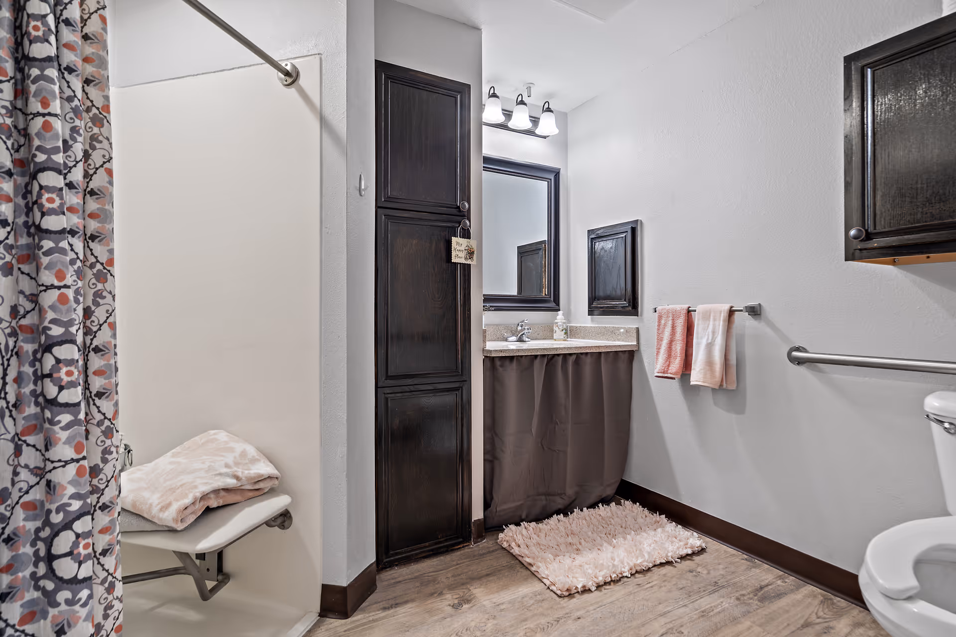 A bathroom with a shower area featuring a patterned curtain and a foldable shower seat with a folded towel on it. There is a dark wood cabinet next to a sink with a mirror above it. Two towels hang on a towel rack next to the sink, and a toilet is partially visible on the right. The floor has a wooden texture with a soft pink rug in front of the sink.