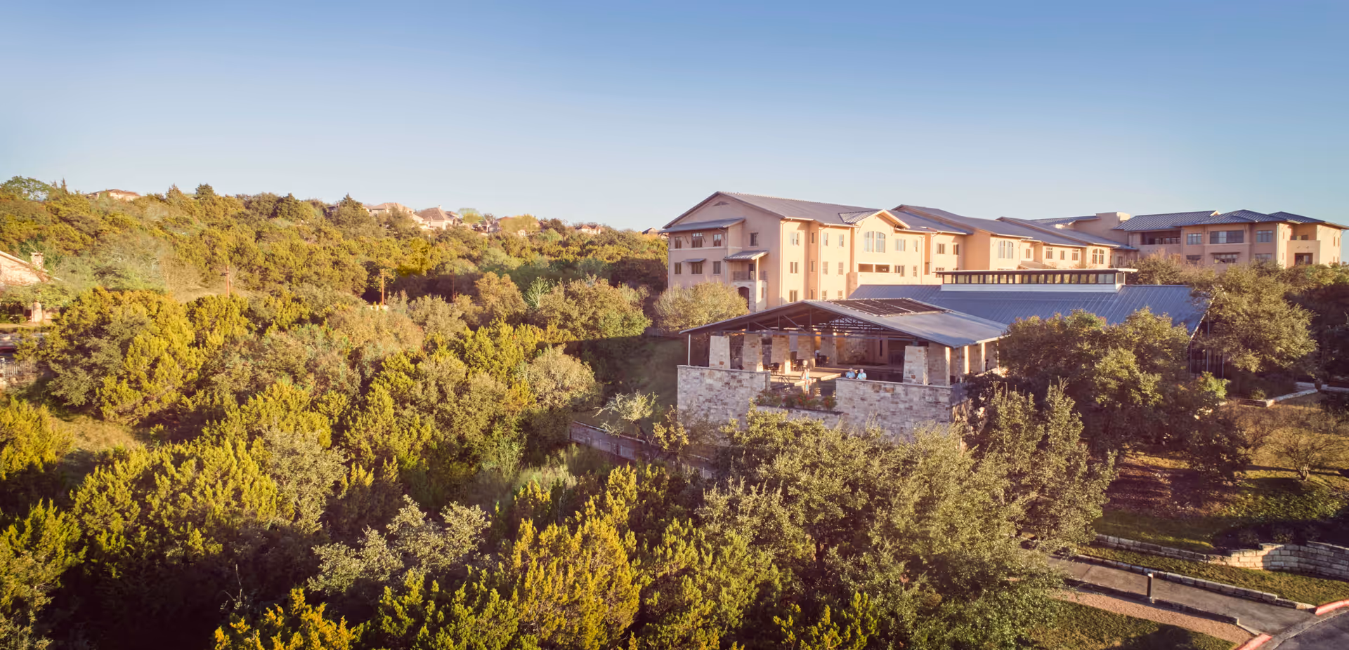 Aerial view of Querencia At Barton Creek senior living facility surrounded by dense green trees and natural landscape under a clear blue sky.