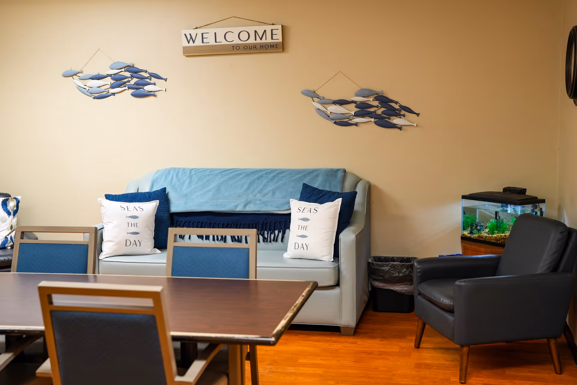 A cozy seating area in a senior living facility with a light blue couch adorned with two white pillows that say 'SEAS THE DAY' and a blue throw blanket. Above the couch, there is a wall decoration of two groups of blue and white fish and a sign that reads 'WELCOME TO OUR HOME'. To the right of the couch is a black armchair next to a small fish tank on a wooden stand. In the foreground, there is a wooden table with blue cushioned chairs.