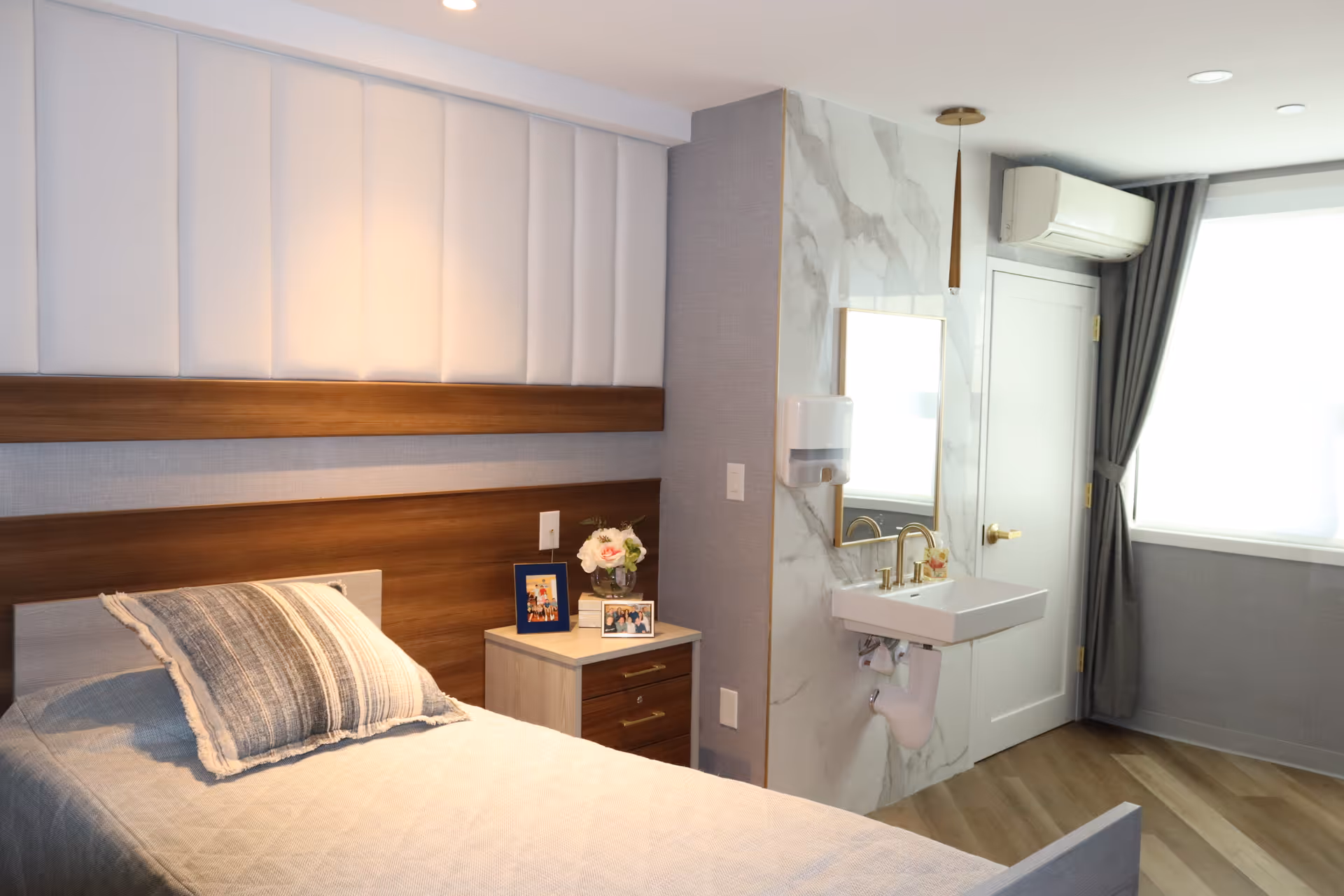 A clean and modern bedroom in a senior living facility featuring a single bed with a striped pillow and beige blanket. Next to the bed is a small nightstand with framed photos and a vase of flowers. On the adjacent wall, there is a small sink with a mirror above it, a soap dispenser, and a door beside a window with gray curtains. The room has wood flooring and soft lighting.