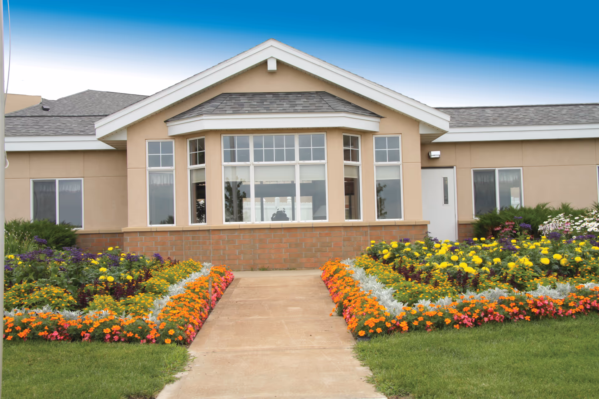 Front entrance of a single-story community building with a central bay window, a walkway and colorful flowerbeds on either side.