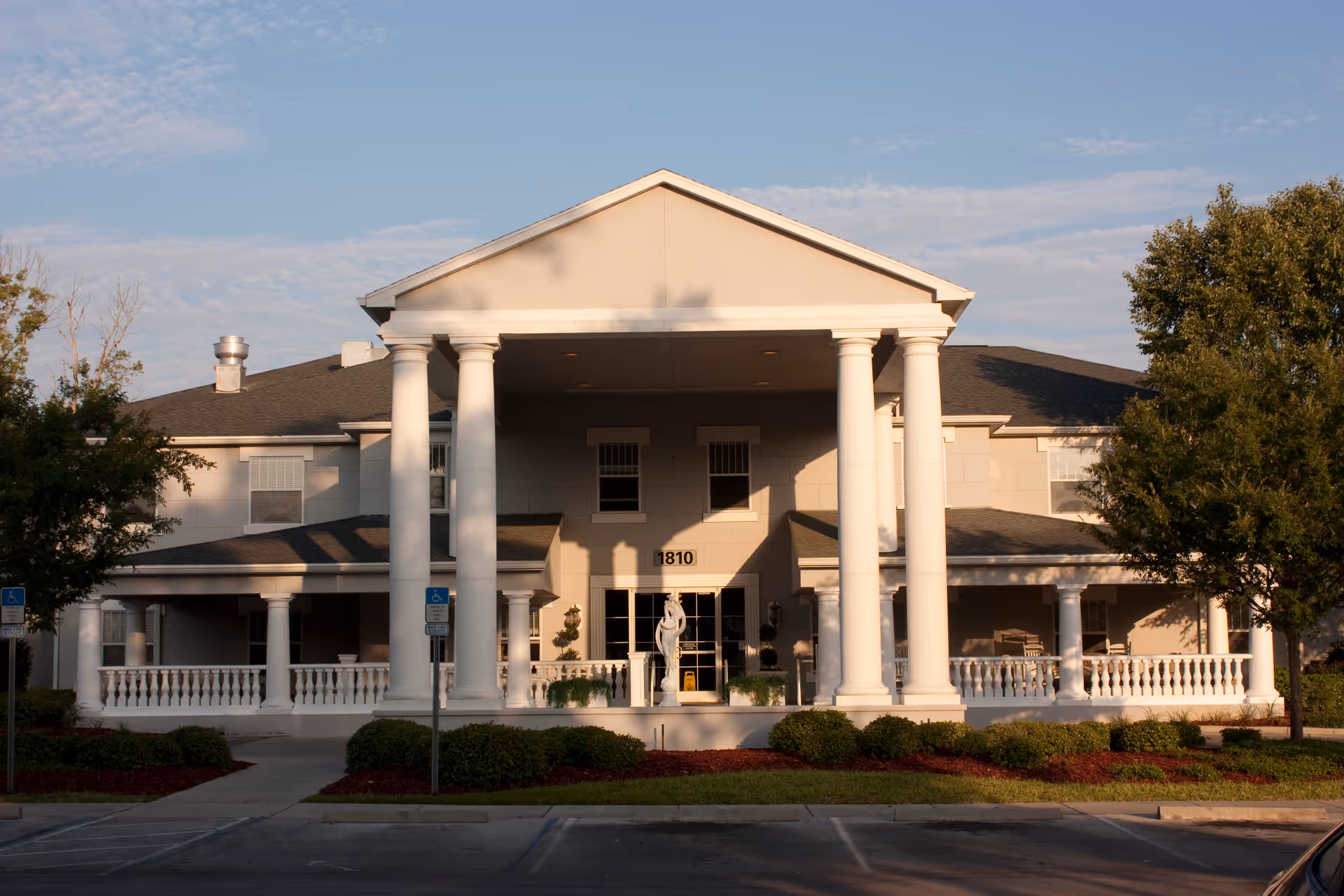 Front exterior view of a two-story building with large white columns and a covered porch area. The building has beige walls, multiple windows, and a sign with the number 1810 above the entrance. There are trees and shrubs around the building and a parking area in front with handicap parking signs.