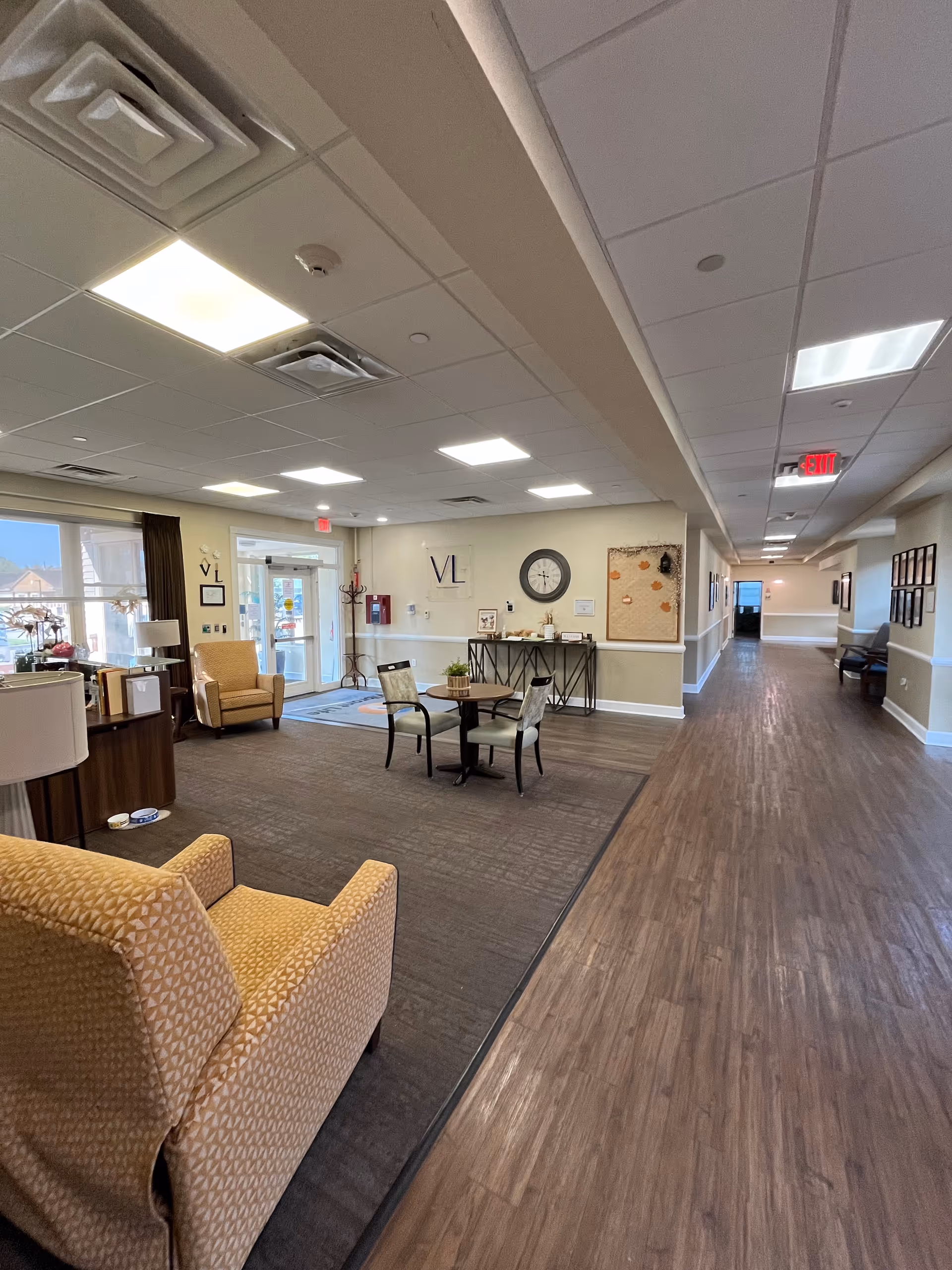 Bright senior living facility lobby with seating, tables, a wall clock, and hallway.