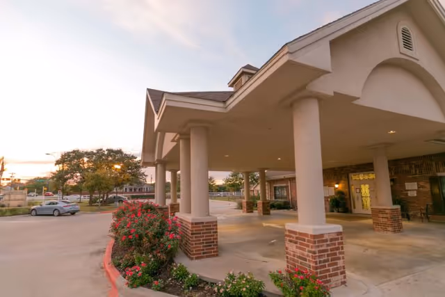 Covered porte-cochère entrance of the Legend Oaks Rehabilitation and Healthcare building with columns, brick planters, and flowering landscaping at dusk.