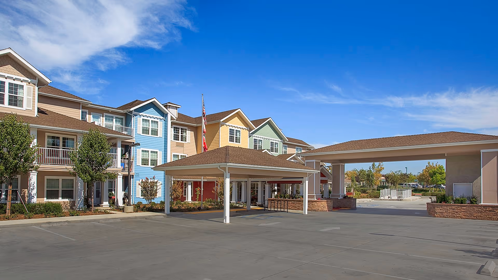 Exterior view of Holiday Chateau at Harveston, showing a multi-story residential building with beige, blue, yellow, and green siding under a blue sky with some clouds. There is a covered entrance area with white pillars and a driveway in front.