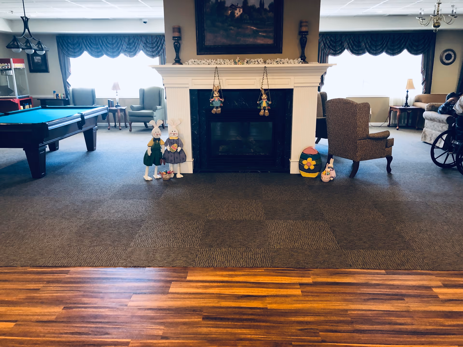 A senior living common room with a central fireplace flanked by chairs and holiday decorations, a pool table to the left, and seating to the right.