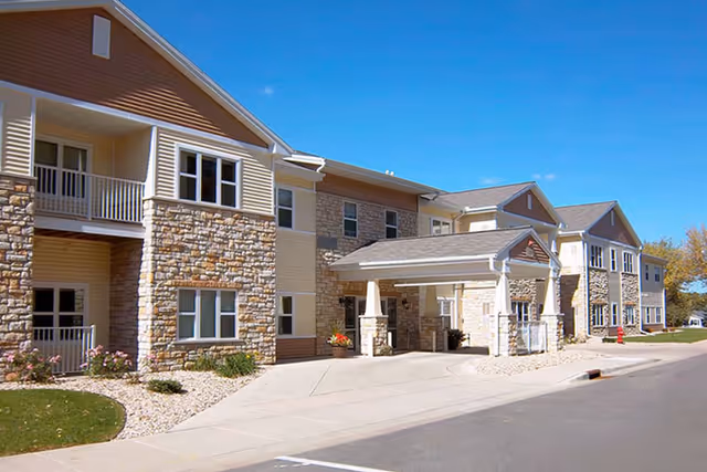 Exterior view of a senior living facility building with stone and beige siding, multiple windows, balconies, and a covered entrance under a clear blue sky.
