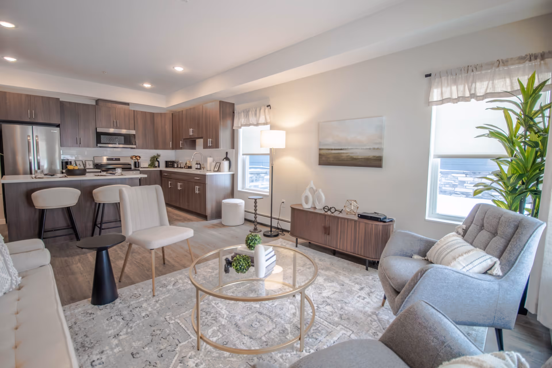 Open-concept living area with gray seating, rug, glass coffee table, and a nearby kitchen.