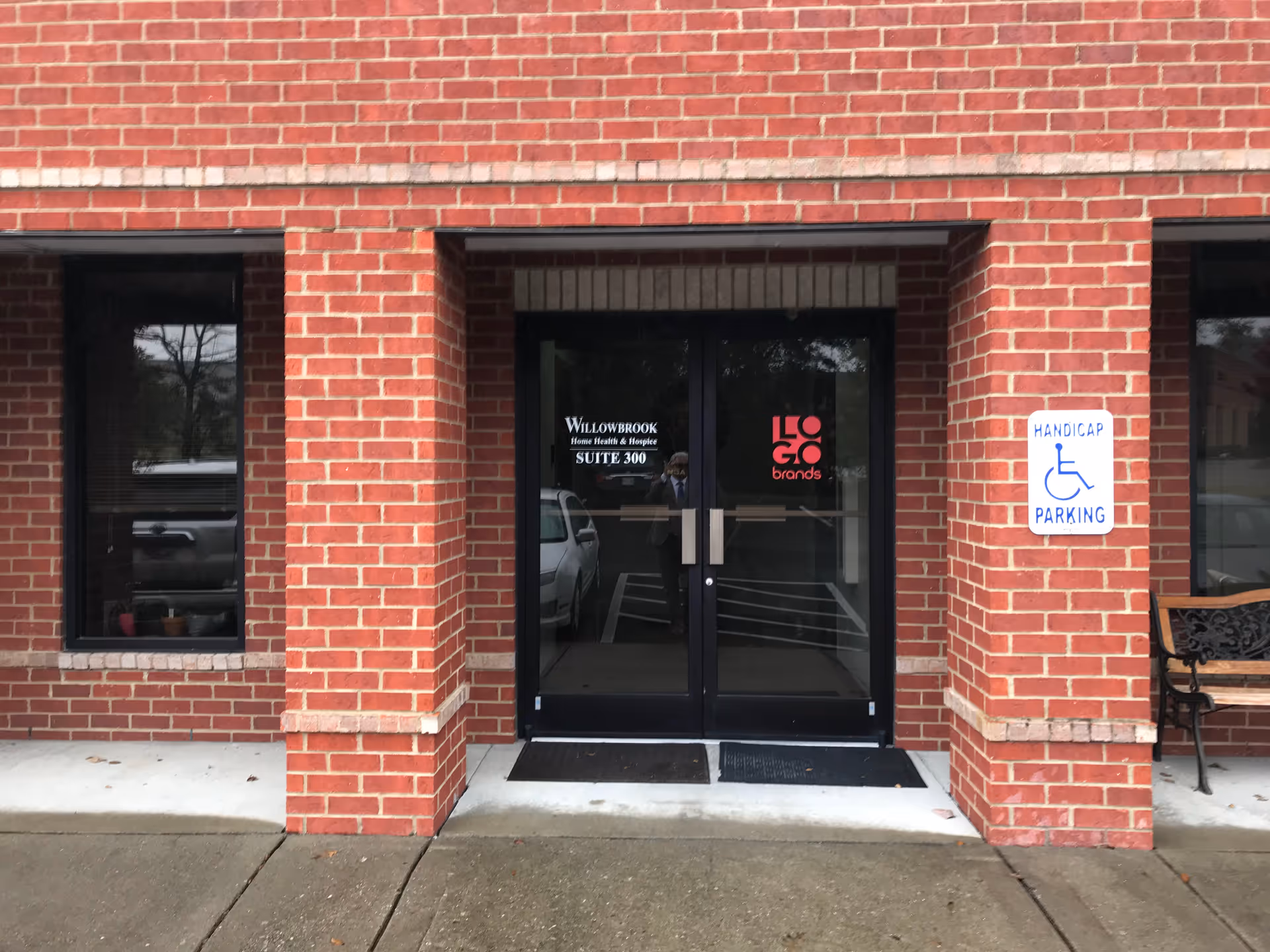 Brick building entrance with glass doors, handicap parking sign, and a bench.