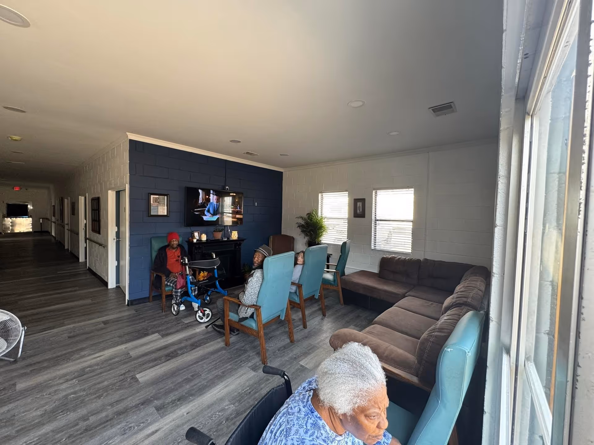 A common area in Carrigg Manor I with several elderly residents seated on chairs and a couch, watching a television mounted on a dark blue accent wall. The room has large windows letting in natural light, wood-look flooring, and a hallway extending to the left with several doors.