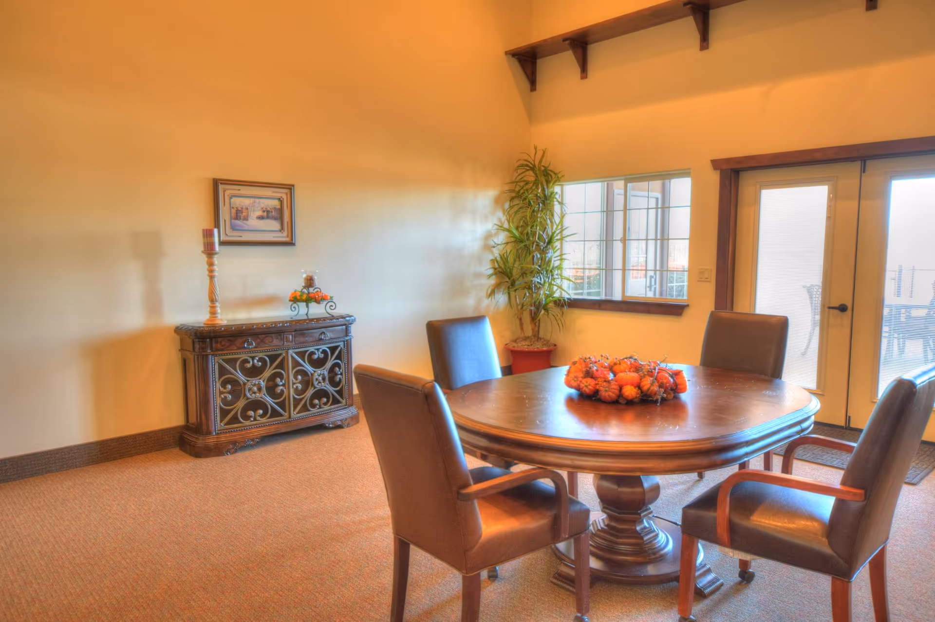 A cozy dining area with a round wooden table surrounded by four brown leather chairs. The table has a centerpiece of small decorative pumpkins. In the background, there is a wooden cabinet with ornate metal doors, a tall candle holder, and a framed picture on the wall. A potted plant is placed near a window and glass doors that lead to an outdoor patio area.