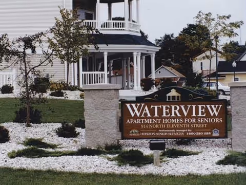 Exterior view of Waterview II senior apartment homes featuring a landscaped area with white rocks, small trees, and a large sign displaying the facility name and address in front of a white building with a porch.