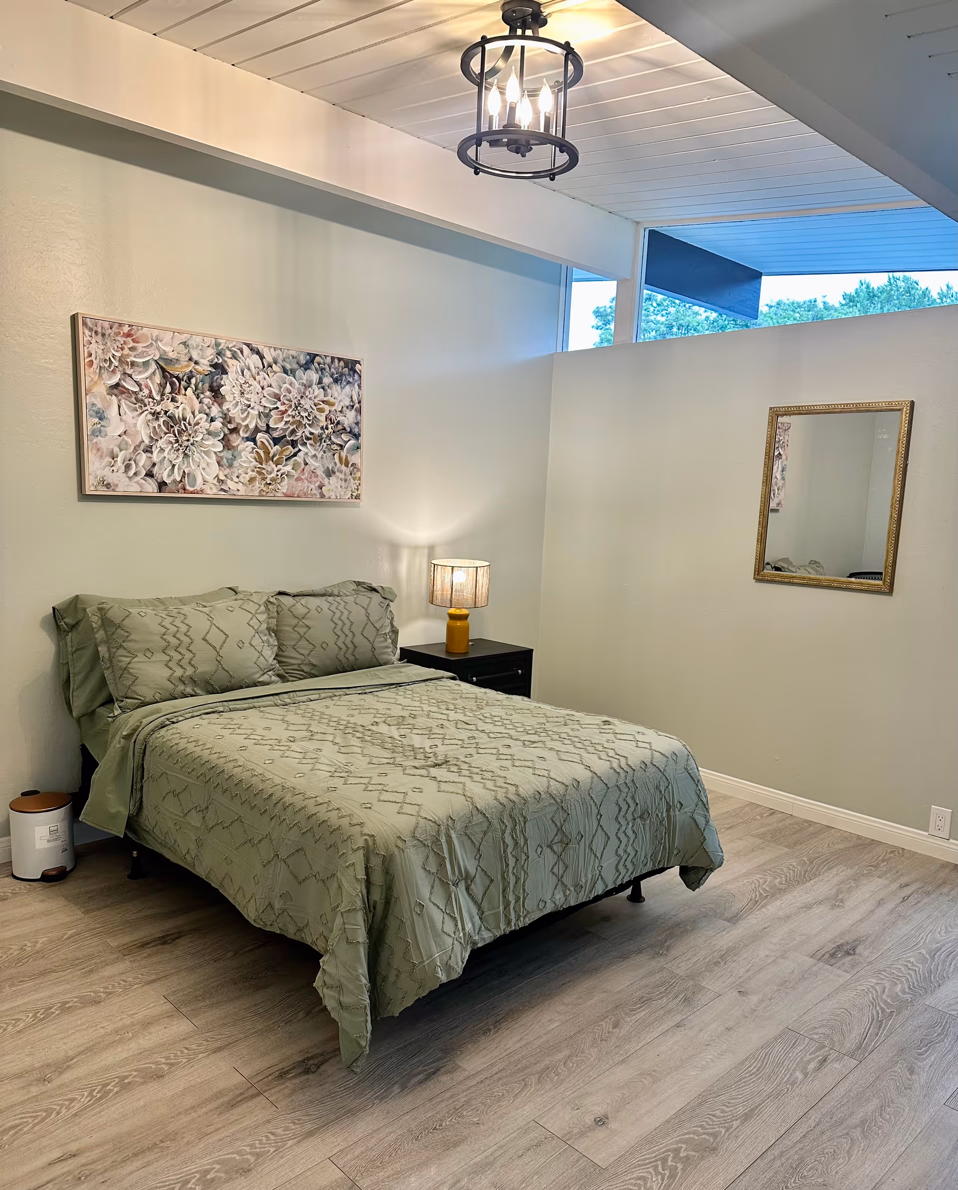 A neatly made bedroom featuring a bed, nightstand, lamp, mirror, and wall art.